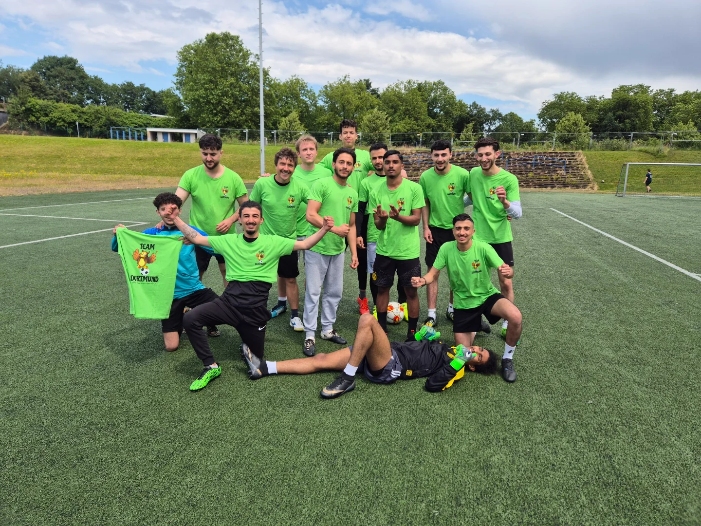 Das Fußballteam der Abendrealschule posiert für ein Mannschaftsfoto auf dem Spielfeld. Ein Spieler hält ein grünes Trikot mit der Aufschrift 'Team Dortmund' hoch.