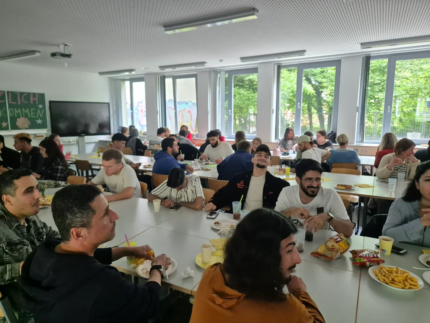 Zahlreiche Studierende und Gäste sitzen beim Sommerfest in einem Klassenraum an Tischen und essen und unterhalten sich.